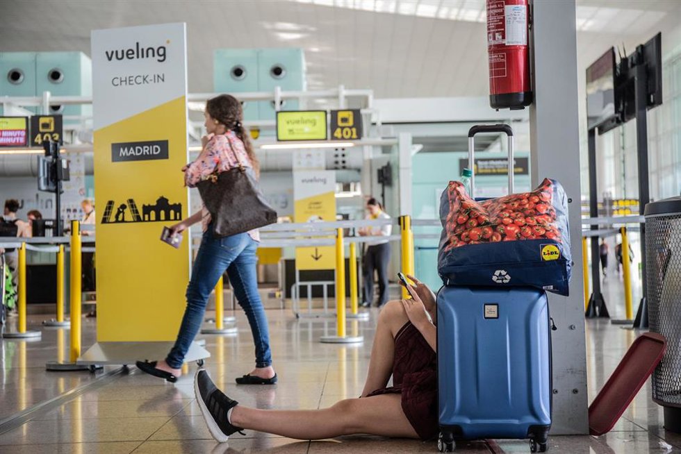 Archivo - Un joven mira su movil sentado junto a su maleta, junto al puesto de Check-in de la compañía Vueling