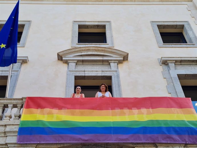 La consellera de Presidencia, Función Pública e Igualdad, Mercedes Garrido, y la directora general de Coordinación, Relaciones con el Parlament, Derechos y Diversidad, Isabel Castro, despliegan la bandera Lgtbi en la sede de la Conselleira de Presidenci