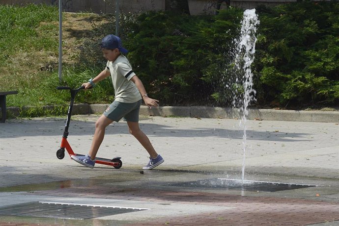 Un niño co un patinete se refresca cerca de una fuente durante la segunda ola de calor de verano en España..