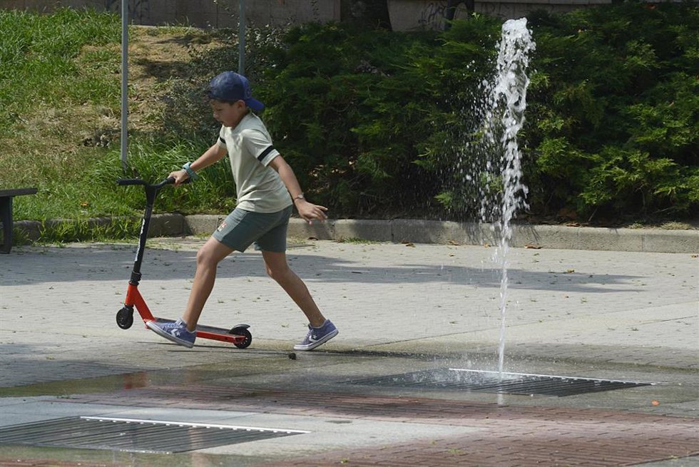Un niño co un patinete se refresca cerca de una fuente durante la segunda ola de calor de verano en España.