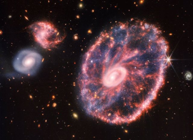 Una gran galaxia rosada moteada que se asemeja a una rueda con un pequeño óvalo interior, con un azul polvoriento en el medio a la derecha, con dos galaxias espirales más pequeñas del mismo tamaño a la izquierda sobre un fondo negro.