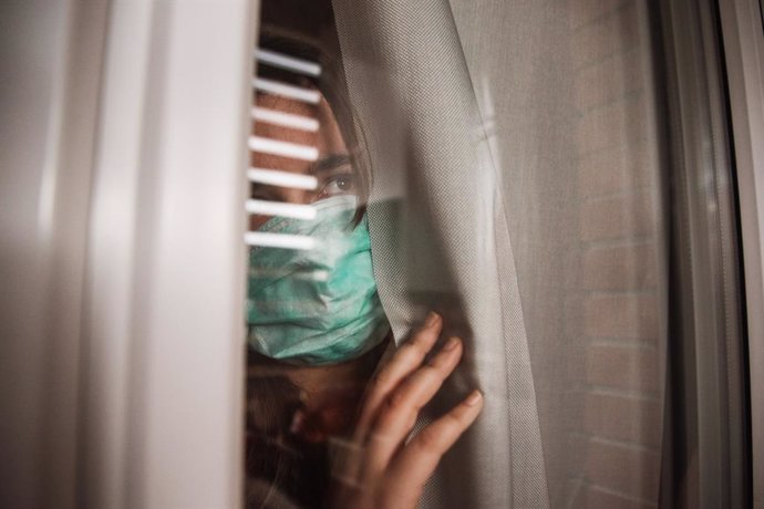 Archivo - Mujer con mascarilla mira con miedo por la ventana.