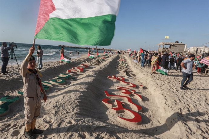 17 August 2022, Palestinian Territories, Gaza City: A Palestinian boy takes part in a commemorative event at the coast of Gaza City as he holds Palestine's national flag while standing next to names carved in the sands for the children killed during the