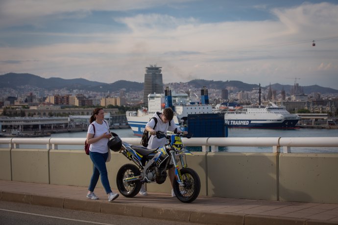Una pareja con una moto camina frente a la terminal de cruceros del Puerto de Barcelona.