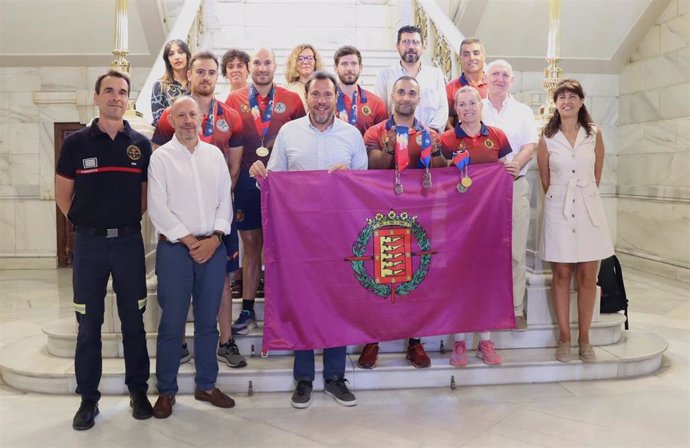 El alcalde de Valladolid, Óscar Puente, varios concejales del equipo de Gobierno y el jefe de Servicio de los Bomberos, Javier Reinoso, posan junto a la bandera de la ciudad y a varios participantes en los Juegos Mundiales para policías y bomberos.