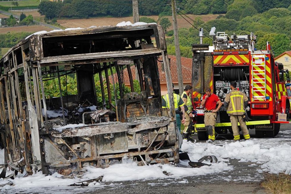 El autobús calcinado junto a varios bomberos durante el dispositivo de extinción del incendio, donde ha fallecido un bombero en prácticas, a 30 de agosto de 2022, en A Cacharela, de la parroquia de Aríns, Santiago de Compostela, A Coruña, Galicia (Españ