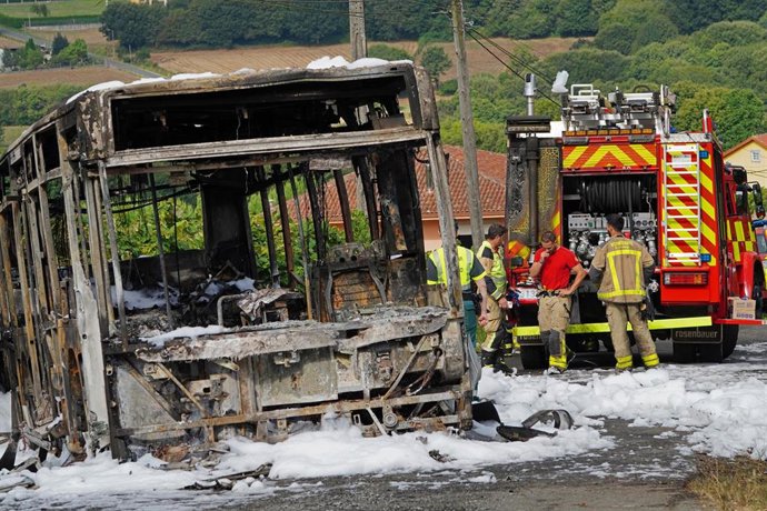 O autobús calcinado xunto a varios bombeiros durante o dispositivo de extinción do incendio, onde faleceu un bombeiro en prácticas, a 30 de agosto de 2022, na Cacharela, da parroquia de Aríns, Santiago de Compostela, A Coruña, Galicia (España)