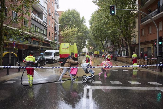 Varias personas cruzan el paso de cebra mientras los servicios de limpieza trabajan en la inundación en la calle Marqués de Vadillo como consecuencia de la rotura de una tubería.