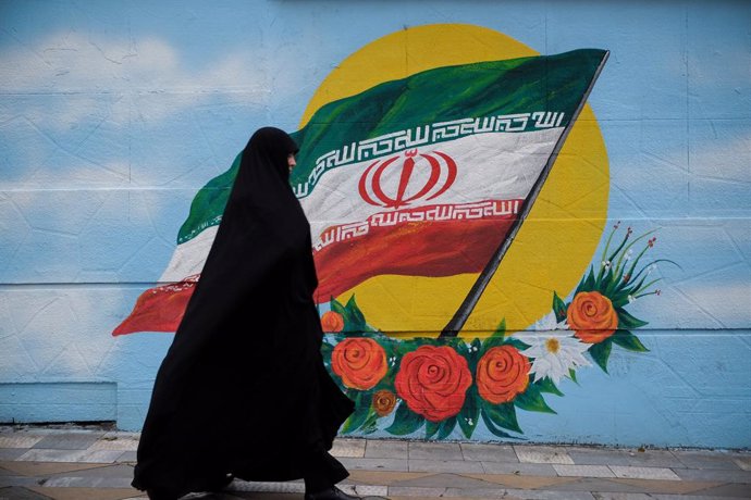 Una mujer con chador camina junto a un mural con la bandera de Irán en Teherán
