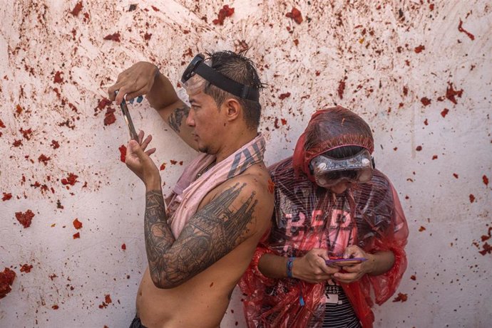 Archivo - Un hombre toma una foto y otro con un chubasquero para no mancharse de tomate, durante la festividad de la Tomatina, a 31 de agosto de 2022, en Buñol, Valencia.