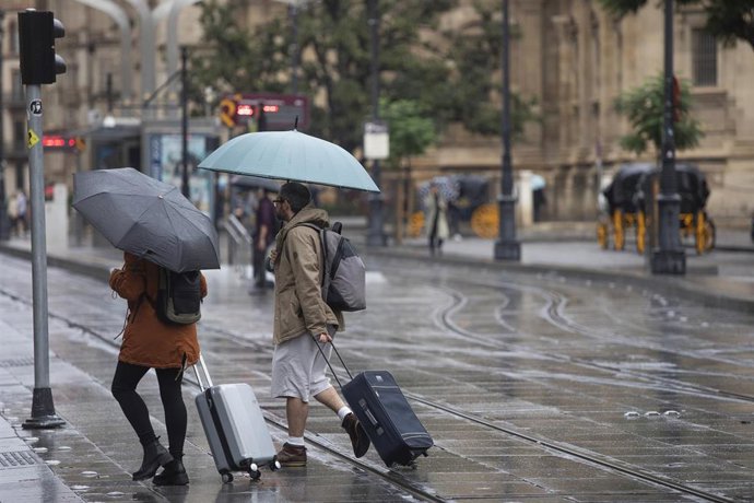 Una pareja con maletas se protegen de la lluvia bajo sus paraguas. A 21 de octubre de 2022, en Sevilla (Andalucía, España). La Agencia Estatal de Meteorología (Aemet) ha ampliado hasta primeras horas de la mañana de este viernes la alerta amarilla por f
