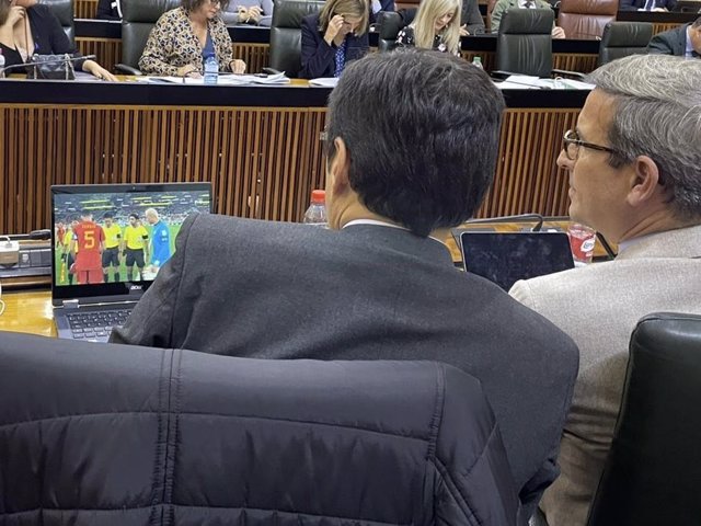 Los consejeros de Justicia, José Antonio Nieto, y de Política Industrial, Jorge Paradela, viendo una imagen del debut de España en el Mundial de Qatar desde su escaño durante el debate de totalidad de los Presupuestos de la Junta para 2023