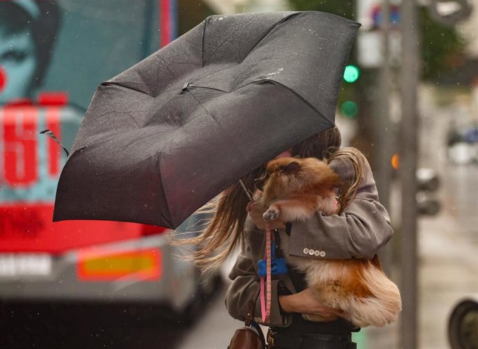 Archivo - Una mujer con un perro intenta controlar y protegerse con un paraguas, en la calle Gran Vía, a 20 de octubre, en Madrid (España). 