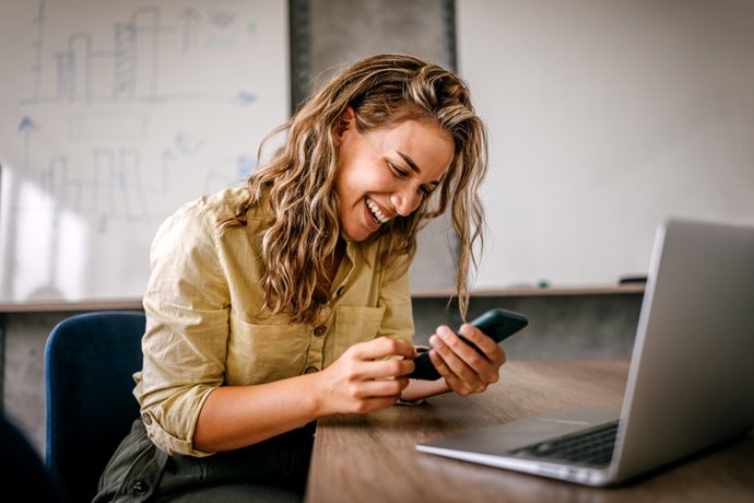 Archivo - Mujeres que usan teléfonos inteligentes y computadoras portátiles riendo