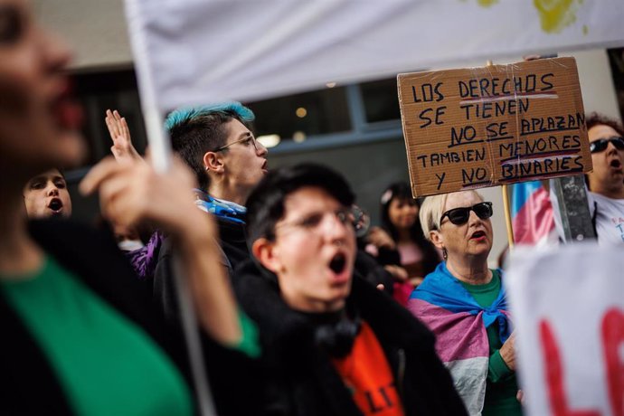 Archivo - Una mujer con una bandera trans sujeta un cartel en el que se lee: 'Los derechos se tienen no se aplazan'
