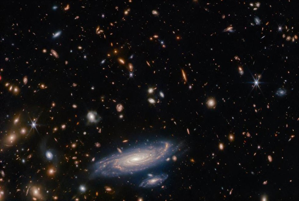Muchas estrellas y galaxias descansan sobre un fondo oscuro, de colores variados, pero sobre todo de tonos anaranjados.