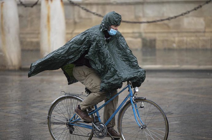 Archivo - Un hombre circula en bicicleta, protegido de la lluvia con un impermeable, junto a la Catedral, en foto de archivo.