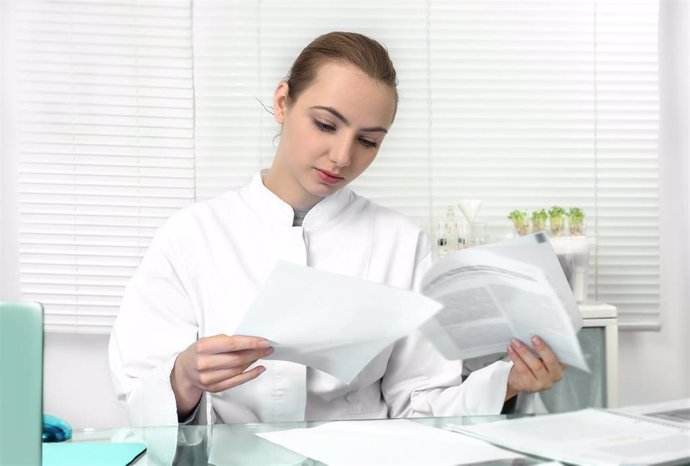 Young Attractive Female Scientist Or Graduate Student In White Coat Reads Scientific Publication. Shallow Dof, Focus On The Face
