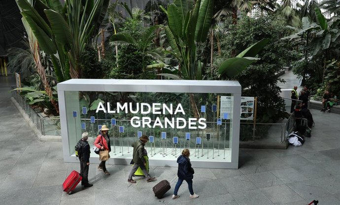 Varias personas caminan frente a la placa de la nueva denominación de la estación Madrid Puerta de Atocha Almudena Grandes como estación Almudena Grandes, en el Jardín Tropical de Atocha-Almudena Grandes.