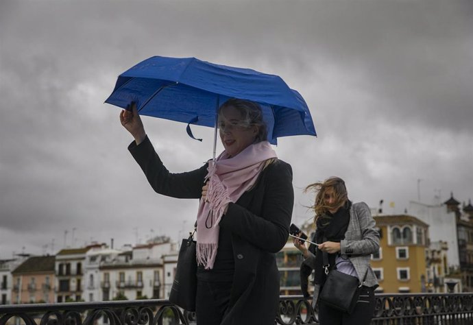 Archivo - Dos mujeres intentan protegerse del viento con su paragüas mientras caminan por el Puente de Isabel II, en Sevilla (Andalucía). 