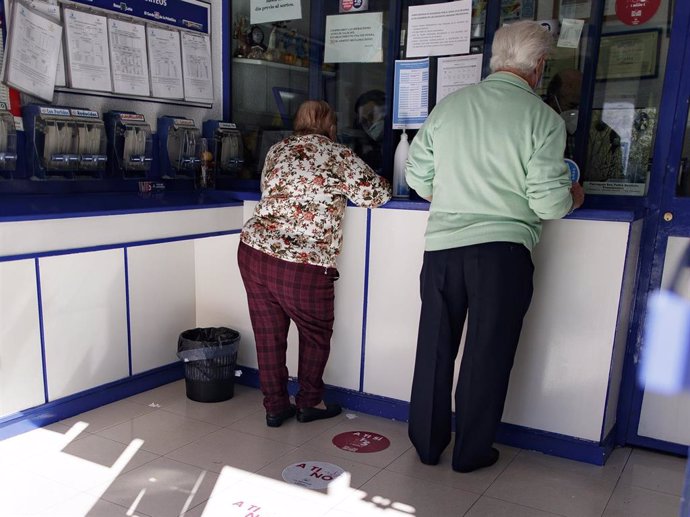 Archivo - Un hombre y una mujer protegidos con mascarillas compran lotería en una Administración
