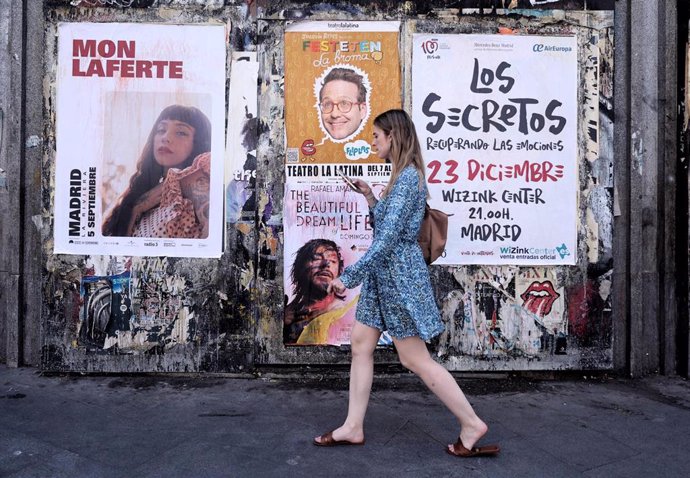 Archivo - Una mujer pasa con su teléfono móvil por varios carteles de conciertos.