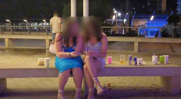 Dos chicas sentadas de noche en una plaza de Magaluf con bebidas alrededor.