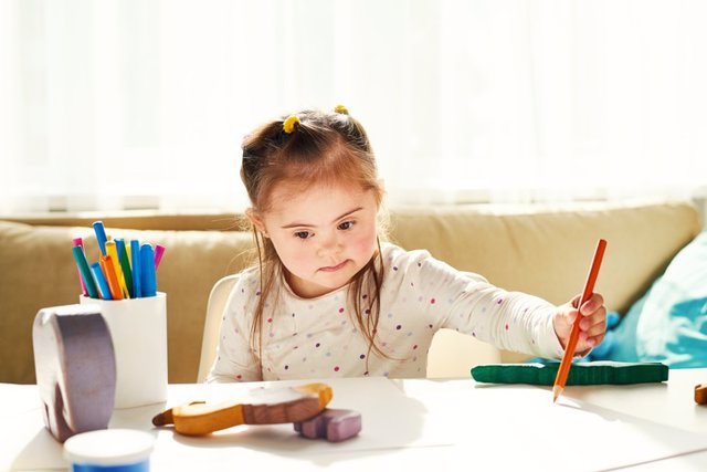 Archivo - Serious cute girl with downs syndrome drawing with orange pencil on paper at desk with toys and art equipments
