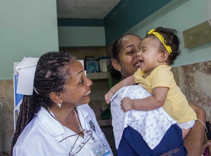Enfermera Sonriendo A Mamá Y Niña En Vacunatorio.