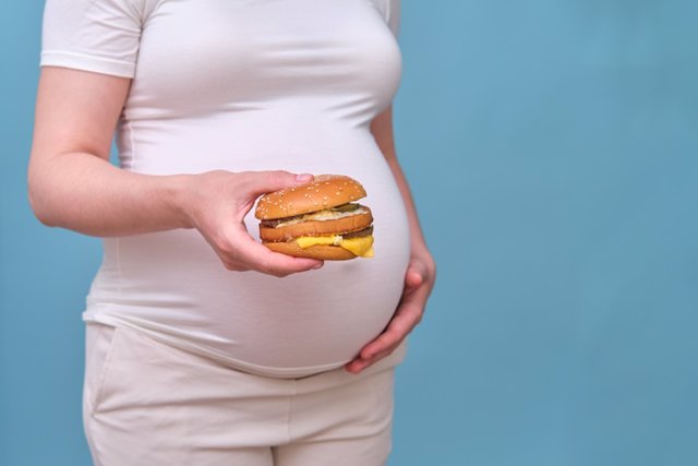 Archivo - Mujer embarazada con hamburguesa sobre fondo azul, primer plano