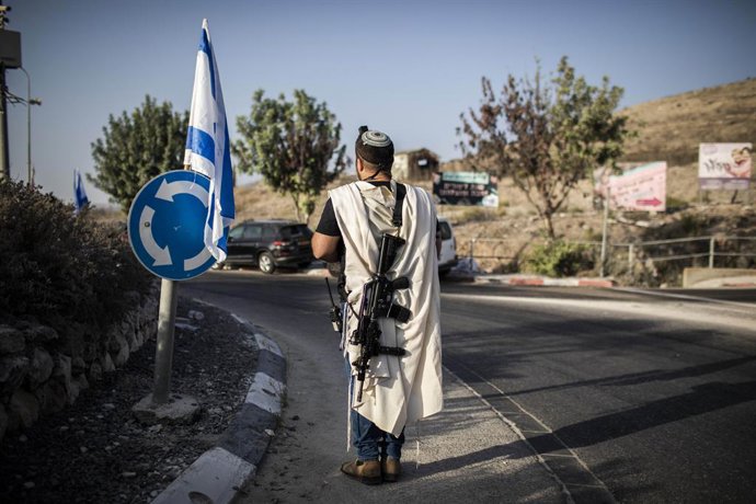 Archivo - 04 October 2022, Palestinian Territories, Nablus: An Israeli right-wing settler carries a weapon by the main entrance to the Palestinian city of Nablus in the northern West Bank while Israeli right-wing settlers pray. Photo: Ilia Yefimovich/dpa