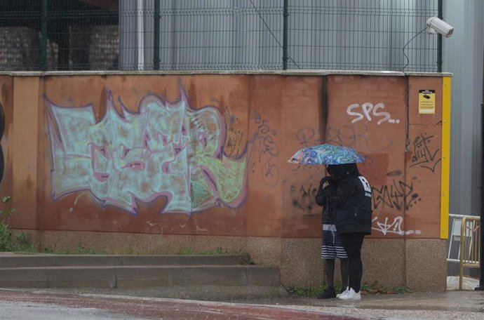Dos migrantes se protegen de la lluvia con un paraguas en las inmediaciones del centro de Emergencia, Acogida y Derivación en el Acuartelamiento 'Primo de Rivera', donde se alojan migrantes procedentes de Canarias, a 19 de enero de 2024, en Alcalá de He