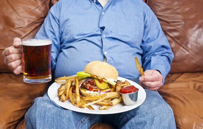 Archivo - Una foto de un hombre con sobrepeso sentado en un viejo sofá con una comida muy abundante y poco saludable en el regazo y una pinta de cerveza en la mano.