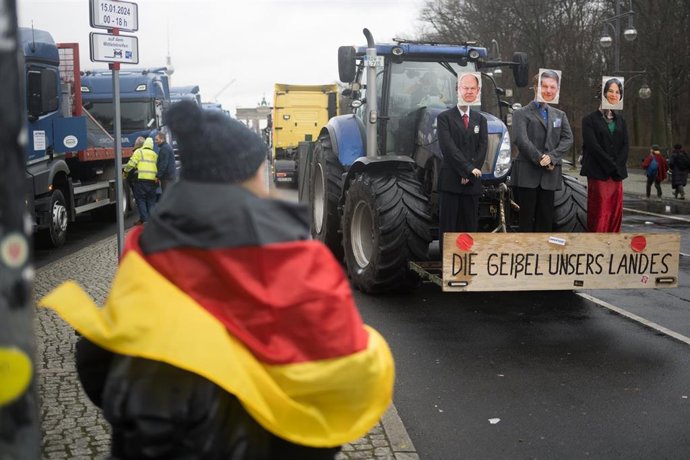 15 January 2024, Berlin: A woman with a German flag stands in front of a tractor on which dolls with the faces of German Chancellor Scholz, Economics Minister Habeck and Foreign Minister Baerbock are mounted with the signature "The scourge of our countr