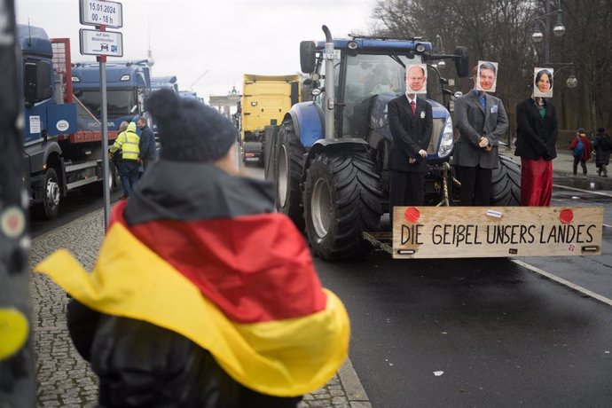 15 January 2024, Berlin: A woman with a German flag stands in front of a tractor on which dolls with the faces of German Chancellor Scholz, Economics Minister Habeck and Foreign Minister Baerbock are mounted with the signature "The scourge of our country"