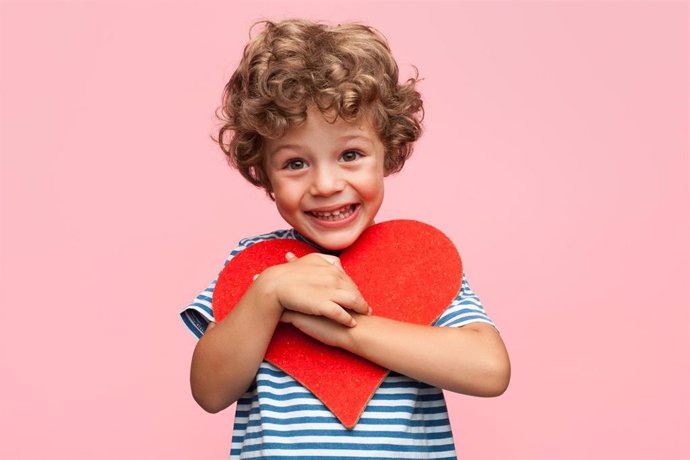 Archivo - Little curly boy holding heart application and laughing at camera on pink background.