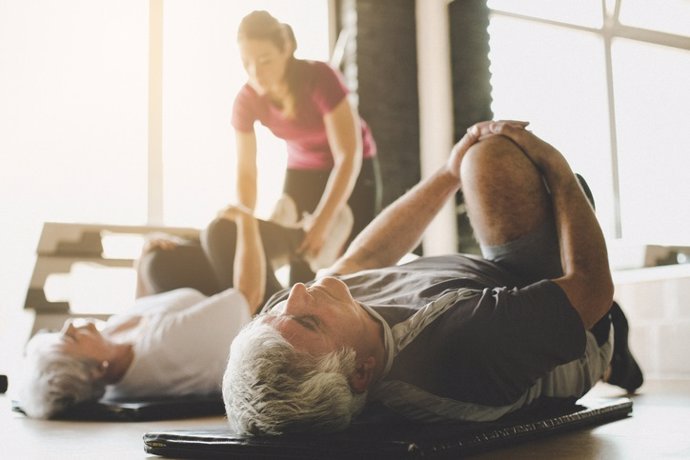 Archivo - Senior couple workout in rehabilitation center. Personal trainer helps elderly couple to do stretching on the floor. Focus on man.