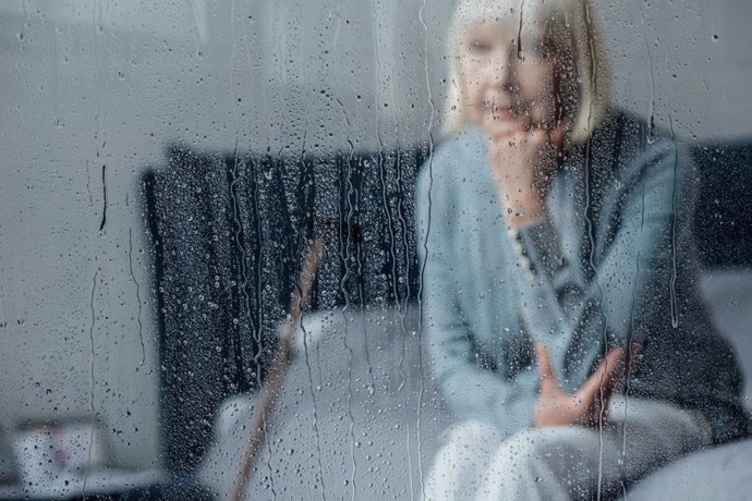 Archivo - Mujer mayor sentada en la cama viendo cómo llueve fuera de casa.