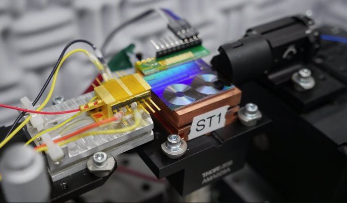 Investigadores del NIST prueban un chip para convertir la luz en señales de microondas. En la foto aparece el chip, que es el panel fluorescente que parece dos pequeños discos de vinilo. La caja dorada  es el láser semiconductor que emite luz al chip