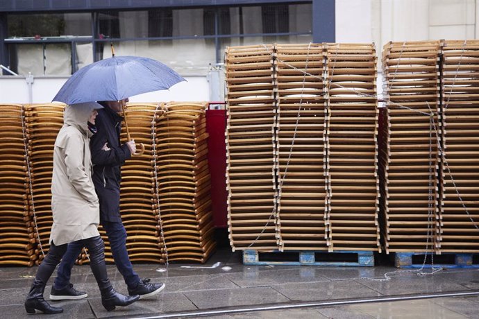 Archivo - Una pareja pasea bajo su paragüa con las sillas apiladas de fondo durante la vuelta de la lluvia a Sevilla en Semana Santa