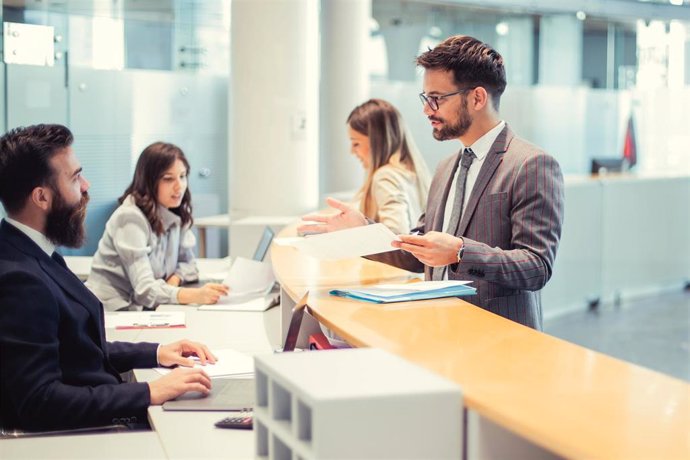 Archivo - A client are standing at the bank counter while the employee is showing him the services available to him. A colleague of him is at the other counter, communicating with a woman.