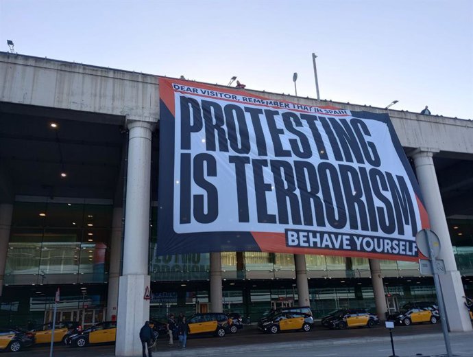 mnium Cultural cuelga una pancarta en el Aeropuerto de Barcelona-El Prat: "Estimado visitante, recuerde que en España protestar es terrorismo. Pórtese bien"