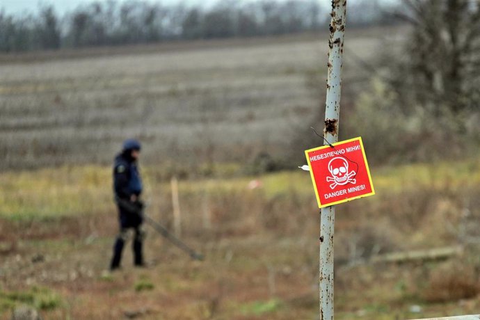 Archivo - Un empleado ucraniano con un detector de minas tras un cartel en el que alerta por la presencia de posibles explosivos