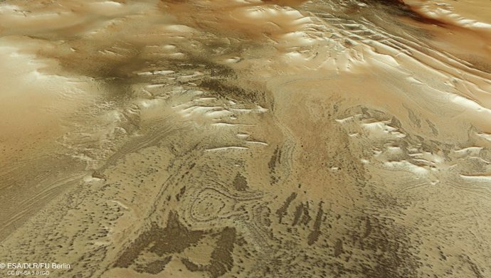 Esta imagen rectangular muestra parte de la superficie marciana como si el espectador estuviera mirando hacia abajo y a través del paisaje, con el suelo irregular y moteado apareciendo en tonos arremolinados de marrón y tostado.