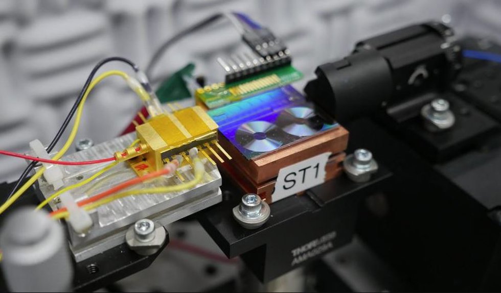 Archivo - Investigadores del NIST prueban un chip para convertir la luz en señales de microondas. En la foto aparece el chip, que es el panel fluorescente que parece dos pequeños discos de vinilo. La caja dorada es el láser semiconductor que emite luz