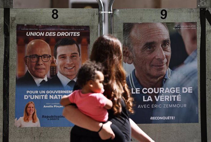 CLICHY-LA-GARENNE, June 30, 2024  -- A mother holding her child walks past posters for legislative elections in Clichy-La-Garenne, near Paris, France, June 30, 2024. The first round of France's snap legislative elections kicks off Sunday in Metropolitan F