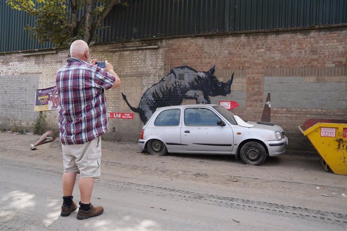 12 August 2024, United Kingdom, London: A man takes a photograph of a new artwork unveiled by Banksy, depicting a rhinoceros which looks as though it is climbing on top of a car Photo: Lucy North/PA Wire/dpa