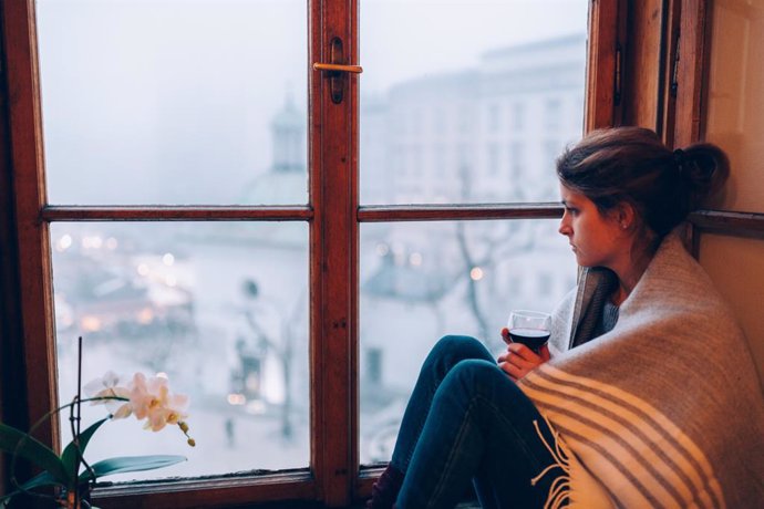 Archivo - Mujer mirando por la ventana en invierno cubierta por una manta.