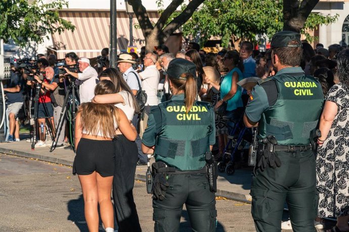 Dos agentes de la Guardia Civil observan a dos chicas abrazadas durante el minuto de silencio por el asesinato de un menor, en la puerta del Ayuntamiento de Mocejón, a 18 de agosto de 2024, en Mocejón, Toledo, Castilla-La Mancha (España). El Ayuntamiento 