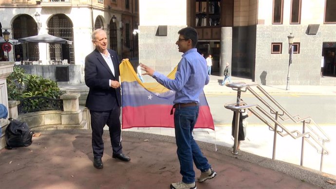 Pedro Gil, coordinador de la Plataforma "Venezolanos en Bilbao", sostiene la bandera de su país ayudado por el exdiputado del PNV Iñaki Anasagasti, natural de Venezuela.